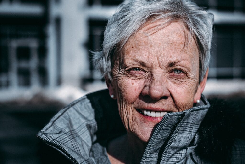 Nahaufnahmeporträt einer älteren Frau mit kurzen grauen Haaren, die warmherzig lächelt und eine graue, karierte Jacke mit hohem Kragen trägt.